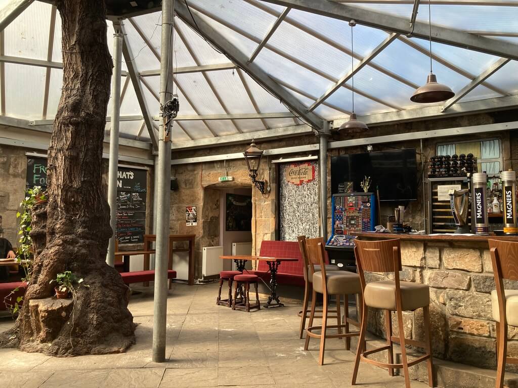 The rustic interior of The Clutha. It has stone walls, a large tree trunk, skylight, and wooden furniture, creating a cozy atmosphere